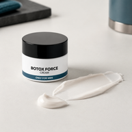 A jar of Botox Force Cream with a black lid and white label, shown on a light gray surface with a smear of the white cream in front.