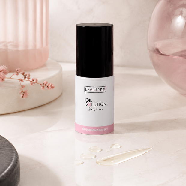 A bottle of Beautyka Oil Solution Nourishing Serum on a light marble surface with a smear of clear serum and soft pink props.