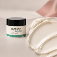 A jar of Overnight Bloom Mask next to a thick swirled smear of the white cream product on a soft beige surface.