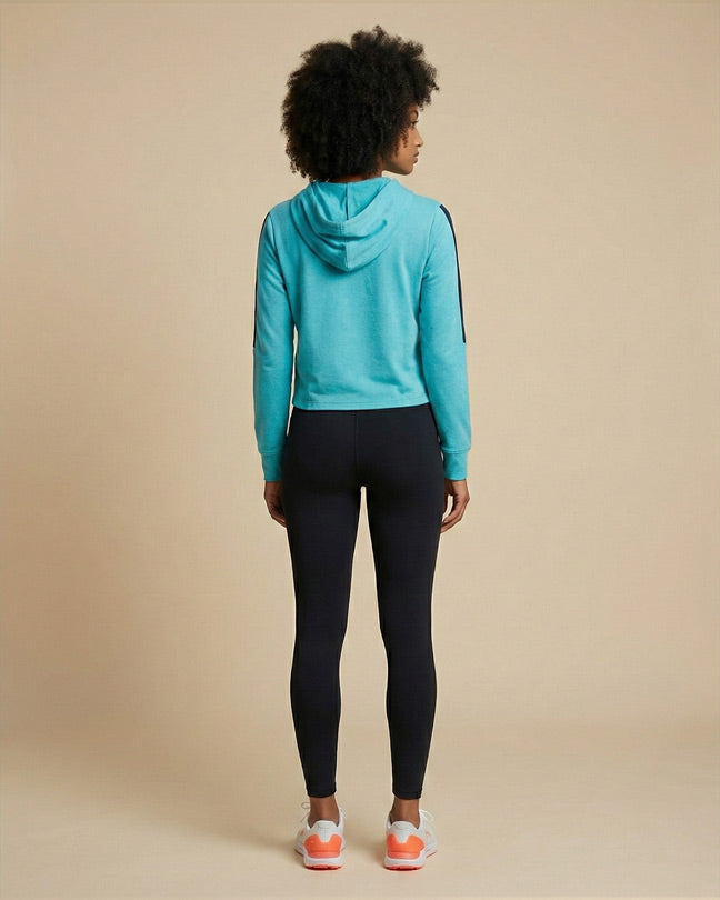 Woman in turquoise hoodie and black leggings, standing indoors, viewed from the back