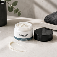 A jar of Reboost Cream for men on a light stone surface with its black lid beside it and a smear of the white cream in front.