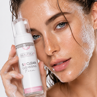 A close-up of a woman with wet hair and foaming cleanser on her face, holding a bottle of BEAUTYKA So Clean Cleansing Foam against a light gray background.