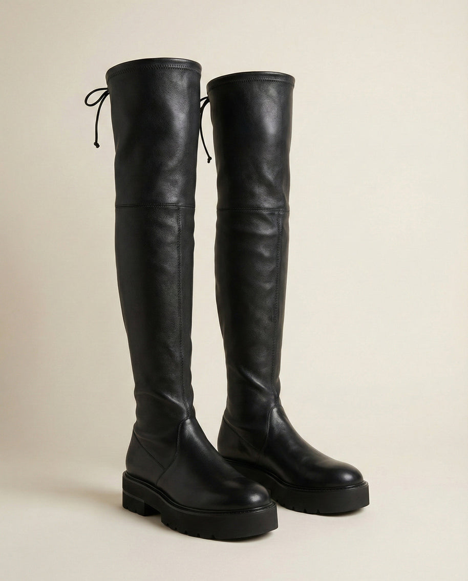 Black leather over-the-knee boots with chunky soles and lace-up detail on a neutral background