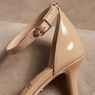 Beige patent leather high heel with ankle strap and gold buckle on neutral background