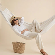 Woman in linen outfit and straw hat relaxing in hammock with woven tote bag and sandals