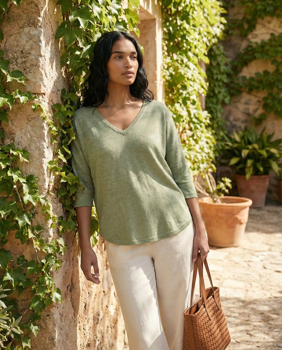 Woman in light green v-neck sweater and white pants holding a brown woven bag outdoors