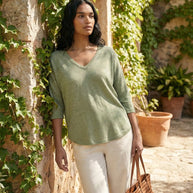 Woman in light green v-neck sweater and white pants holding a brown woven bag outdoors