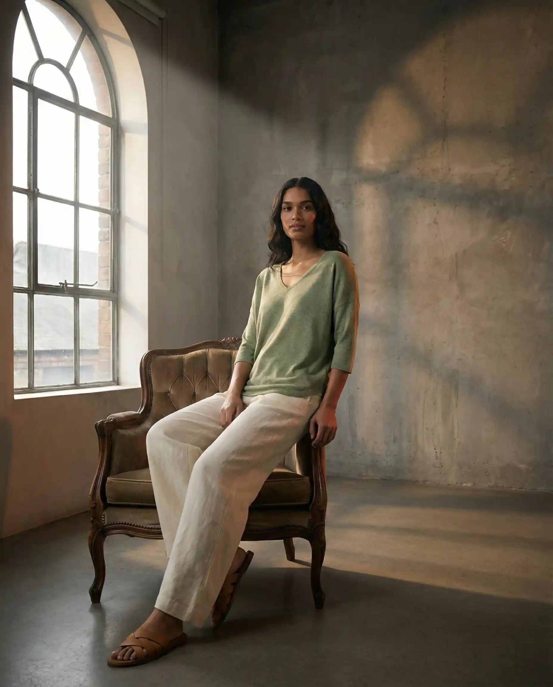 Woman in green top and white pants sitting on vintage chair by arched window in sunlit room