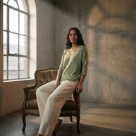 Woman in green top and white pants sitting on vintage chair by arched window in sunlit room