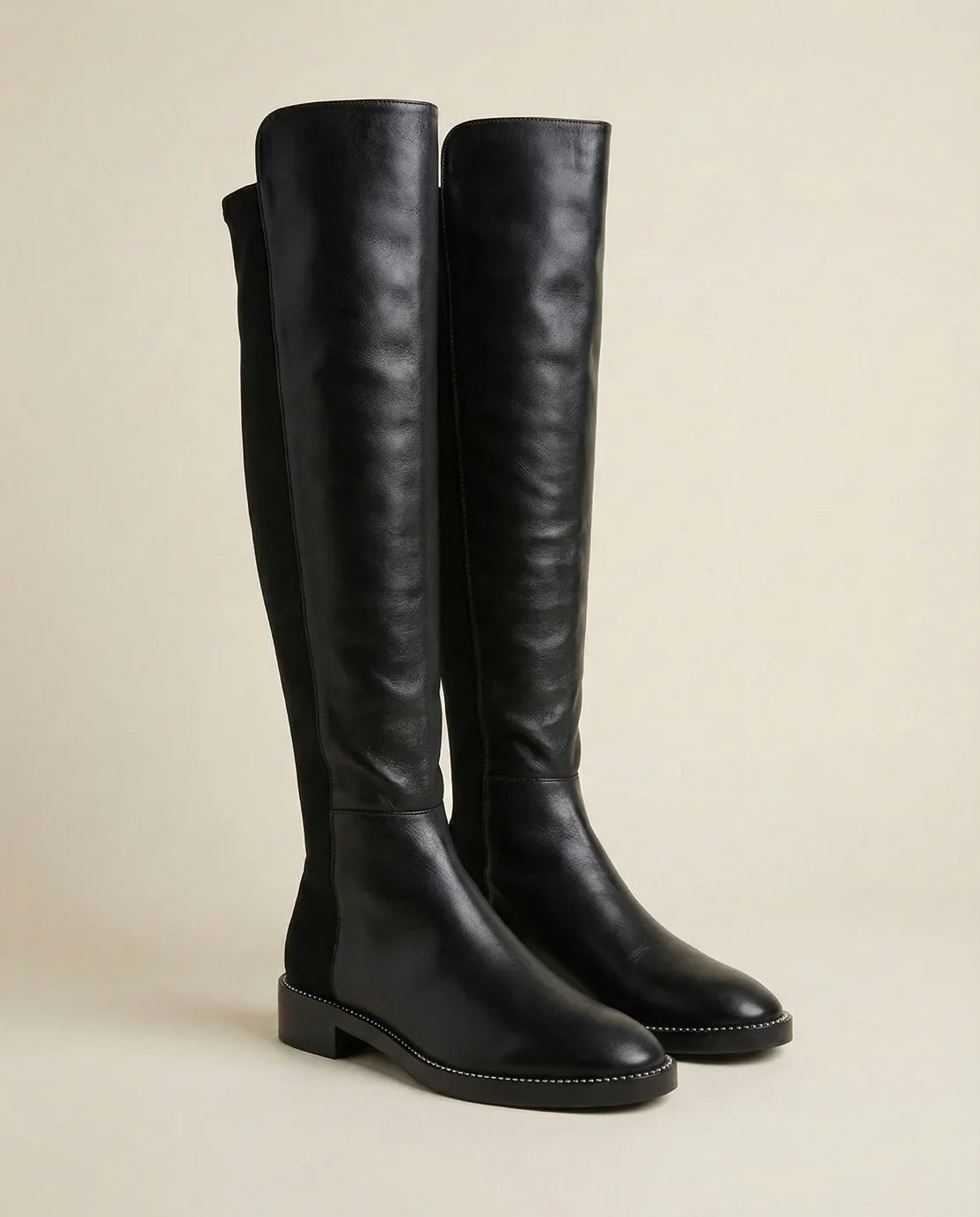 Black knee-high leather boots with low heels and stitching detail on neutral background