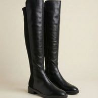 Black knee-high leather boots with low heels and stitching detail on neutral background