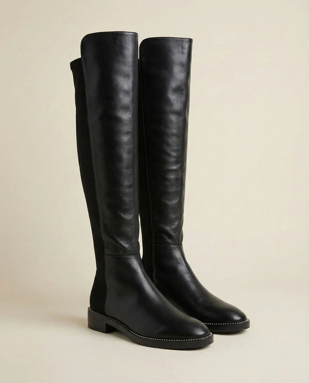 Black knee-high leather boots with low heels and stitching detail on neutral background