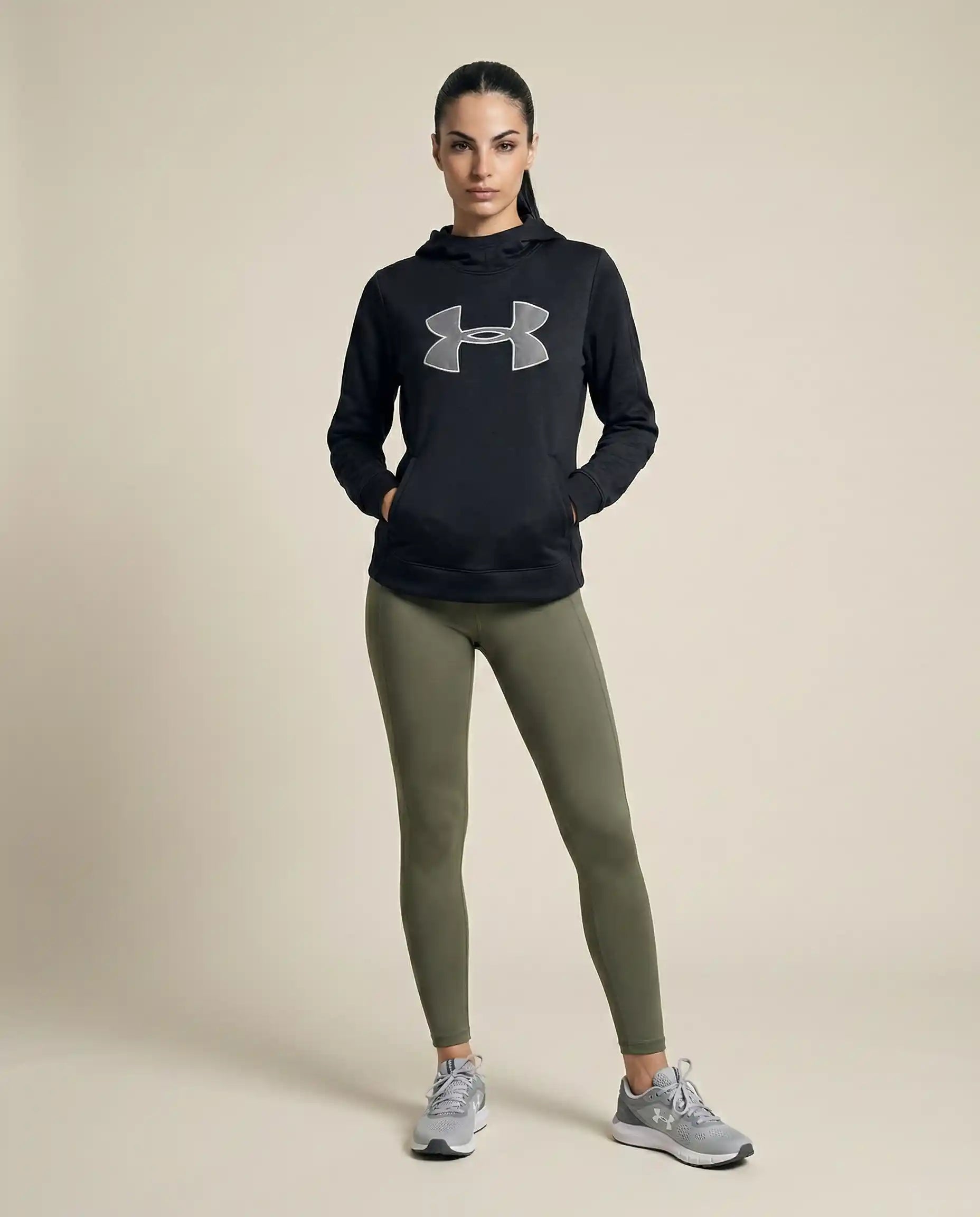 Woman wearing black athletic hoodie, olive green leggings, and gray sneakers, studio background
