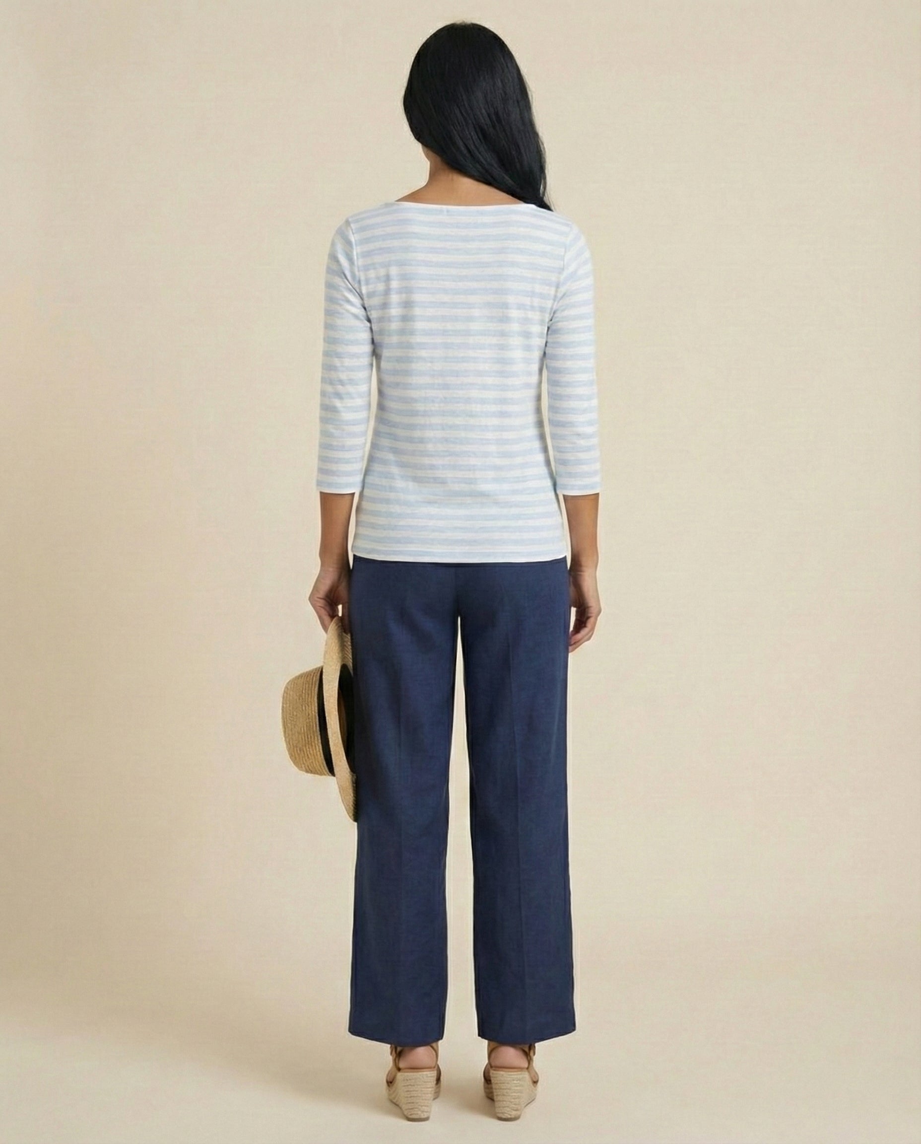 Woman in striped top and blue pants holding a straw hat, back view, neutral background