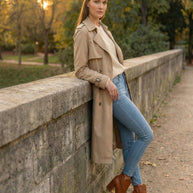 Woman in beige trench coat, blue jeans, and brown boots standing by stone wall in park
