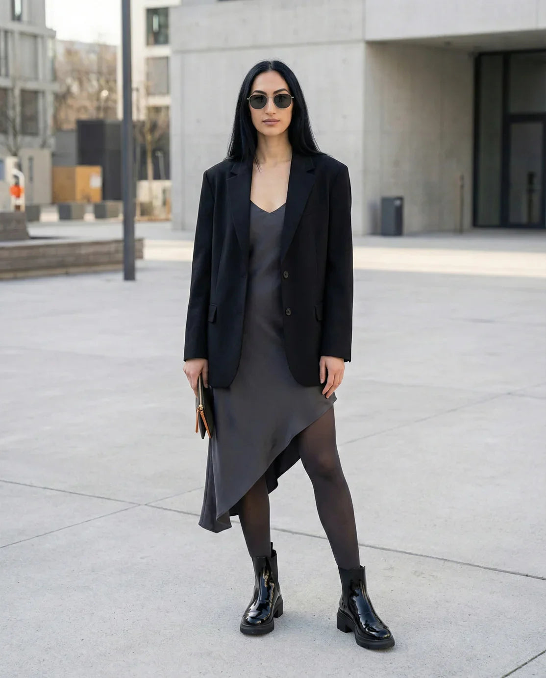 Woman in a black blazer, asymmetrical slip dress, ankle boots, and sunglasses outdoors