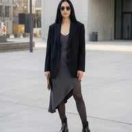 Woman in a black blazer, asymmetrical slip dress, ankle boots, and sunglasses outdoors