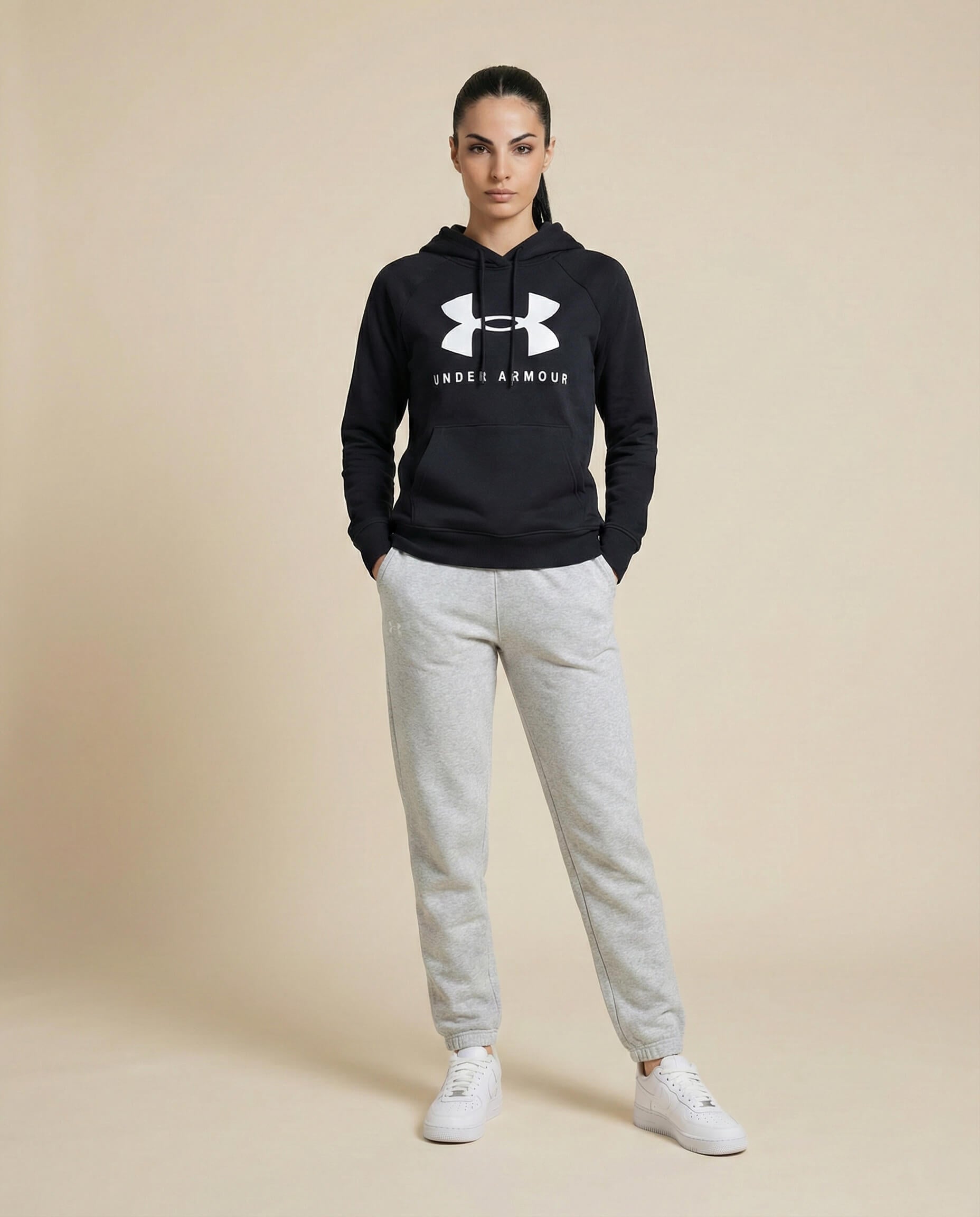 Woman in black Under Armour hoodie, gray joggers, and white sneakers on beige background