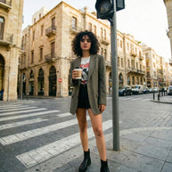 Woman in a blazer, graphic tee, black boots, and shorts holding coffee on a city street crosswalk
