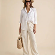 Woman in white linen shirt and beige pants, straw hat, sandals, and woven tote bag