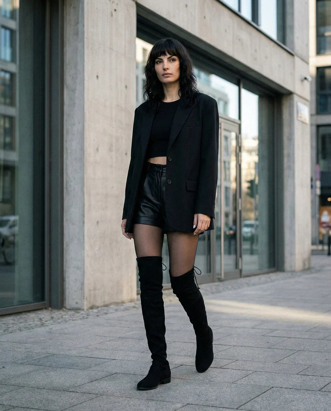 Woman in black blazer, crop top, leather shorts, sheer tights, and over-the-knee boots walking in city