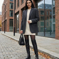 Woman wearing black blazer, knit sweater dress, over-the-knee boots, and holding handbag on city street