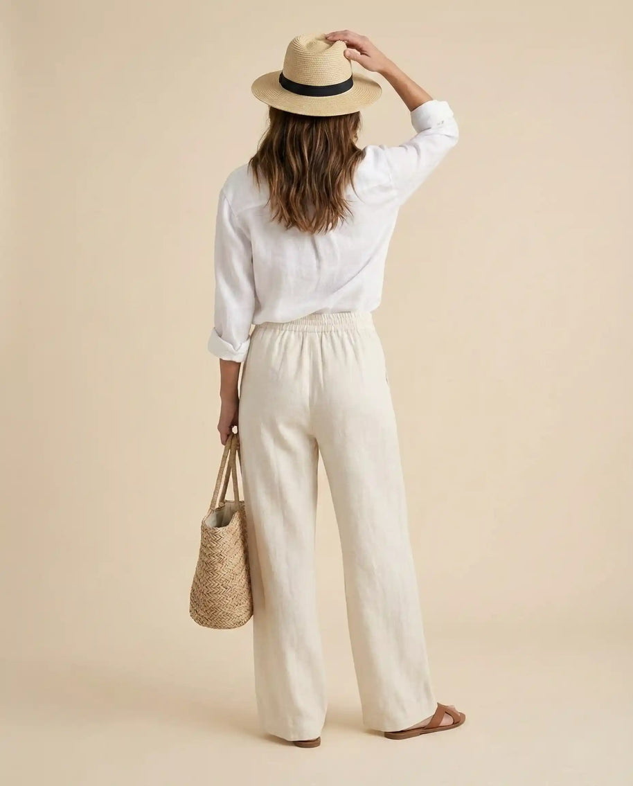 Woman in white linen outfit, straw hat, and sandals holding woven tote bag, beige background
