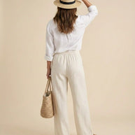 Woman in white linen outfit, straw hat, and sandals holding woven tote bag, beige background