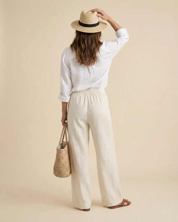 Woman in white linen outfit, straw hat, and sandals holding woven tote bag, beige background