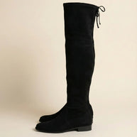 Black suede over-the-knee boots with low heels and tie detail on a neutral background