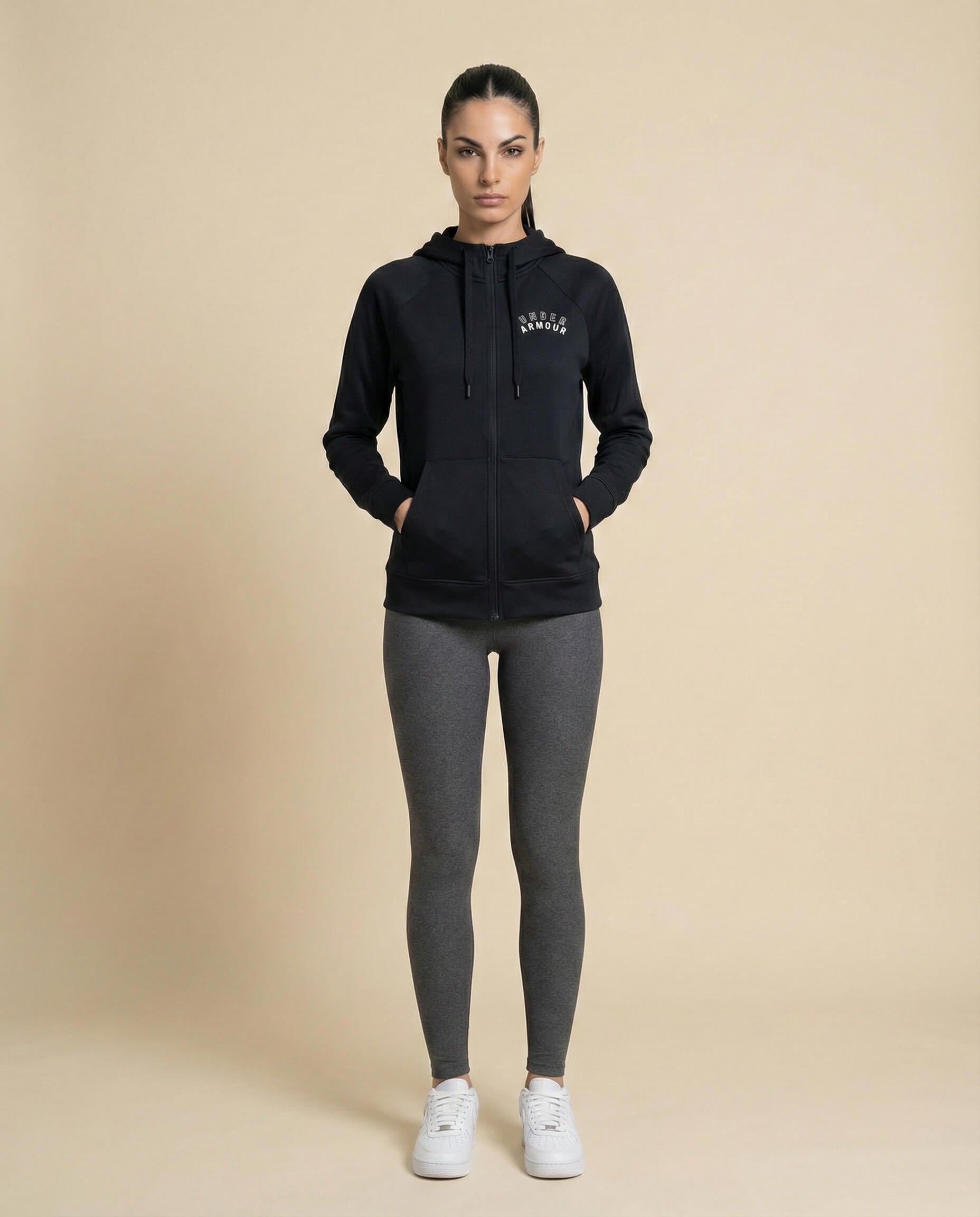 Woman in black zip-up hoodie, gray leggings, and white sneakers, standing on beige background