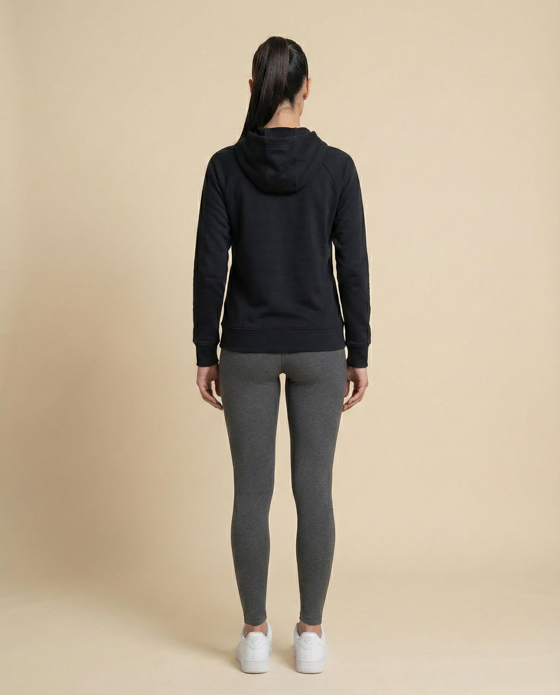Woman in black hoodie and gray leggings, back view, on beige background, casual outfit