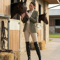 Woman in equestrian outfit stroking a horse outside stable with hay and tack visible