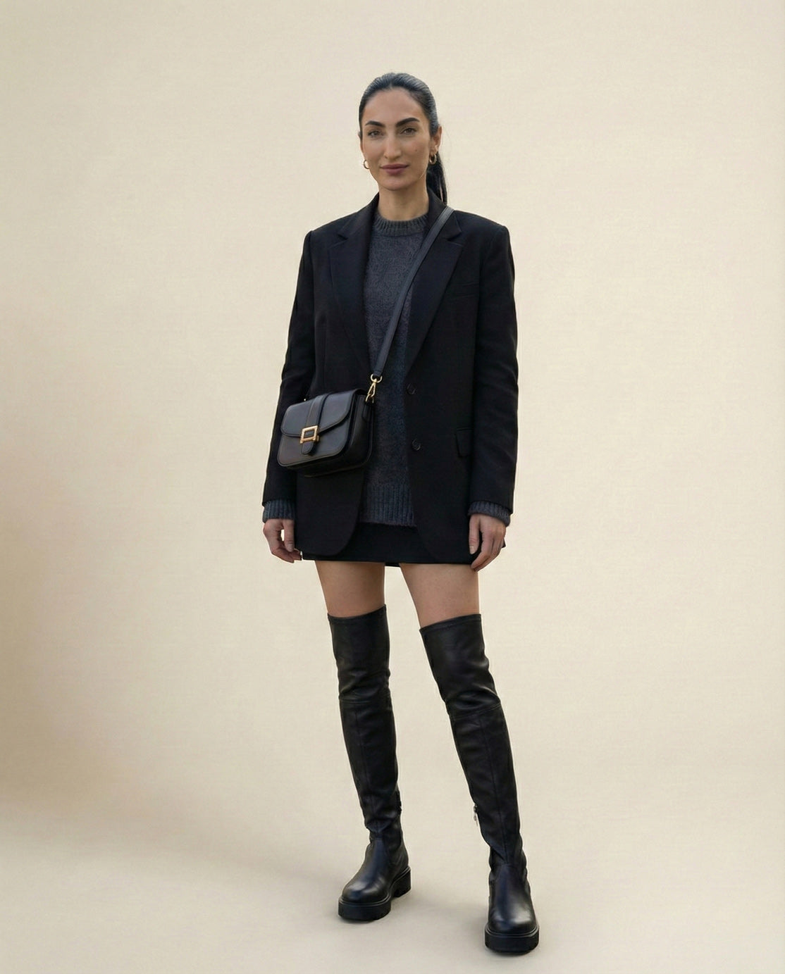 Woman in black blazer, gray sweater, mini skirt, and over-the-knee black boots with crossbody bag