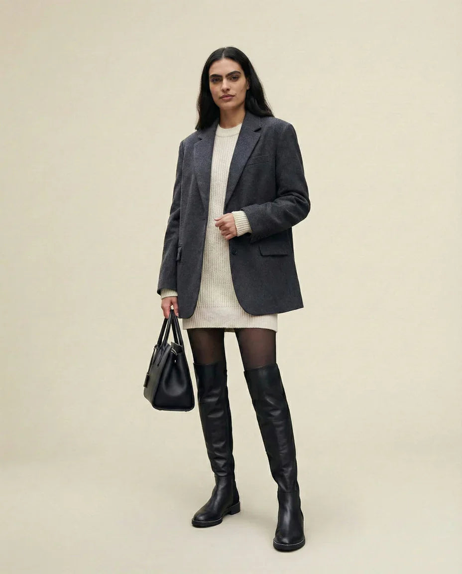 Woman in gray blazer, cream sweater dress, black over-the-knee boots, and holding black handbag