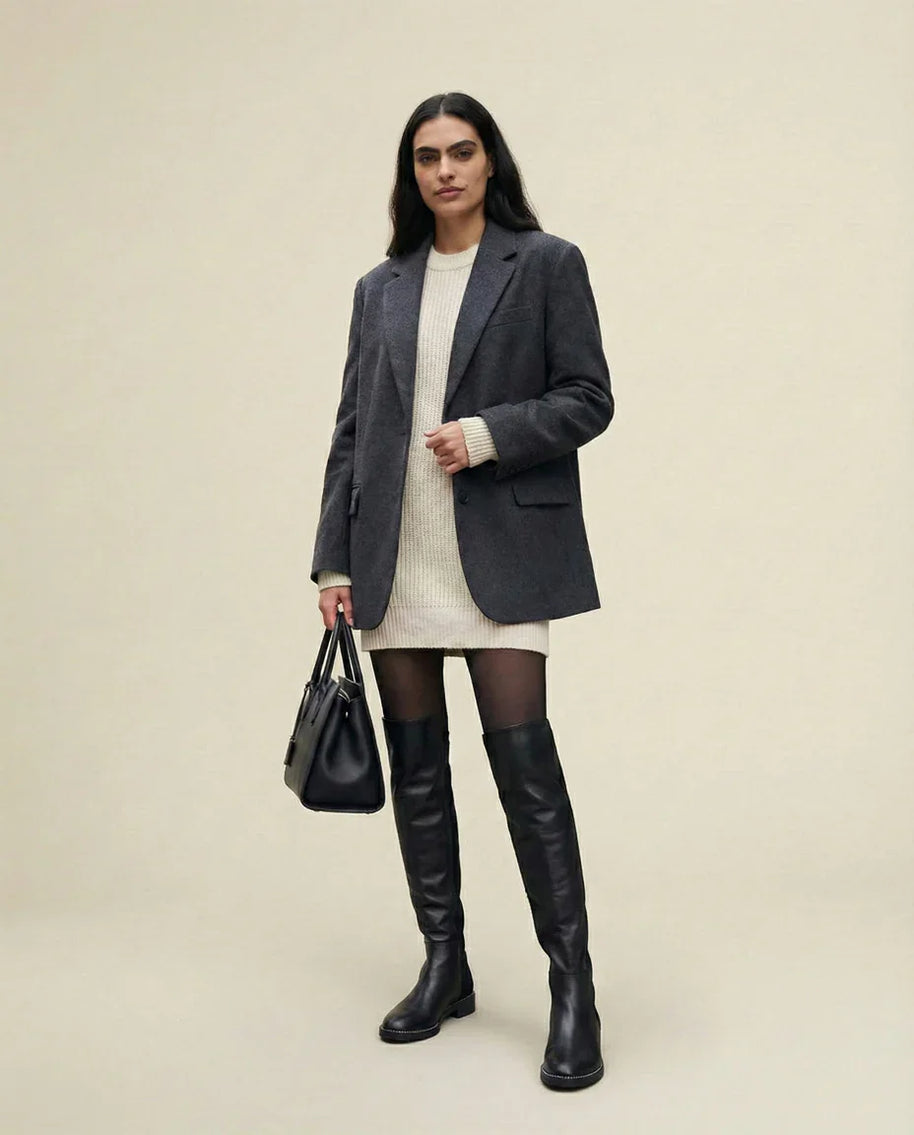 Woman in gray blazer, cream sweater dress, black over-the-knee boots, and holding black handbag