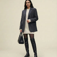 Woman in gray blazer, cream sweater dress, black over-the-knee boots, and holding black handbag