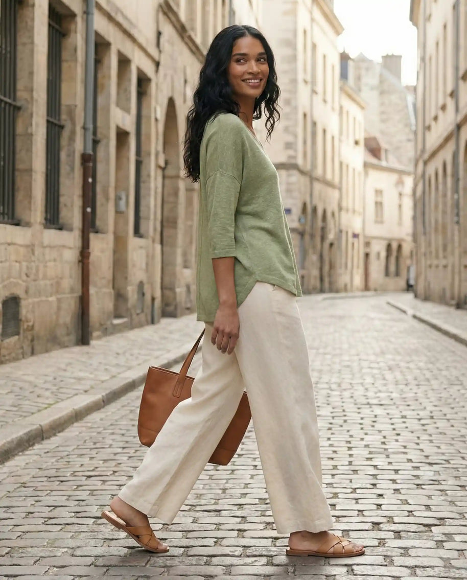 Woman in green top and beige wide-leg pants walking on a cobblestone street with a brown tote