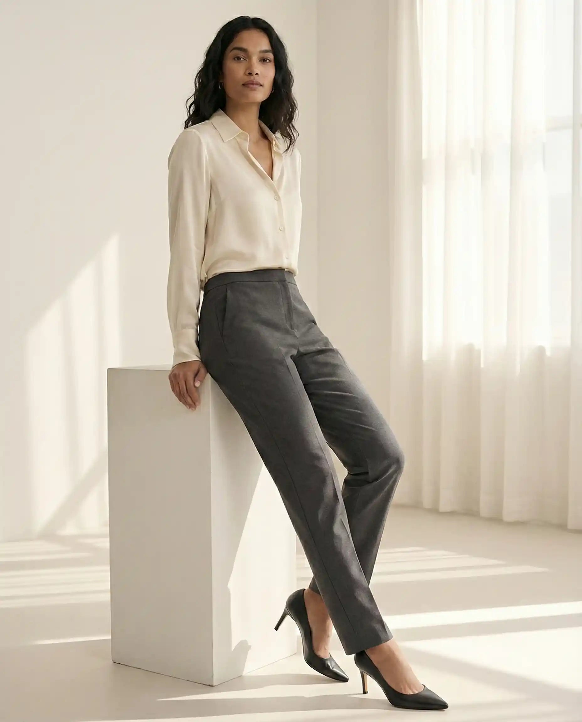 Woman in cream blouse and gray dress pants with black heels in sunlit minimalist room