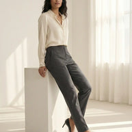 Woman in cream blouse and gray dress pants with black heels in sunlit minimalist room