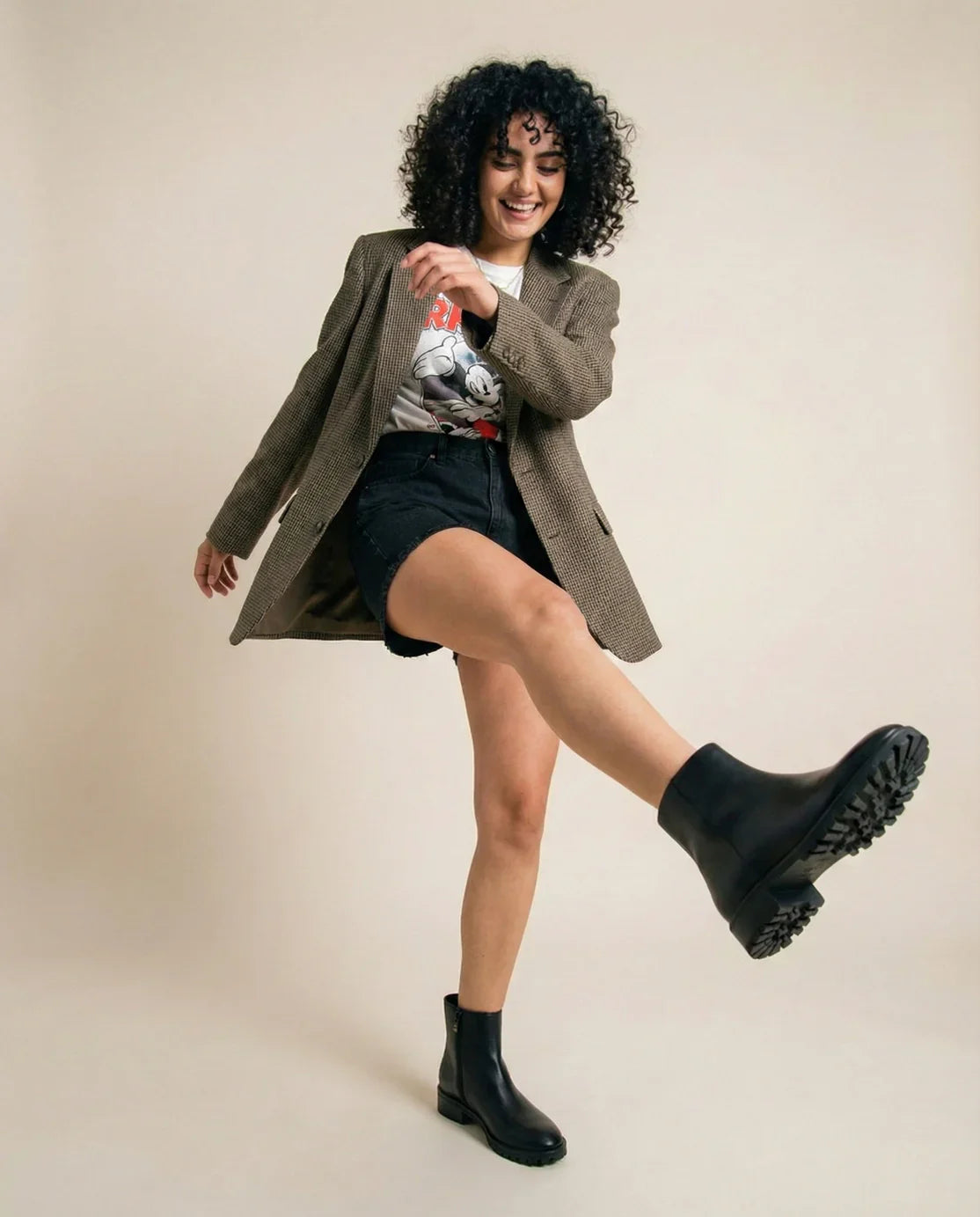 Woman in casual outfit with graphic tee, blazer, black shorts, and chunky black boots posing playfully.