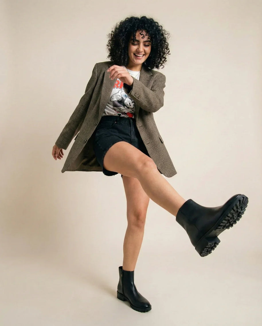 Woman in casual outfit with graphic tee, blazer, black shorts, and chunky black boots posing playfully.