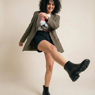 Woman in casual outfit with graphic tee, blazer, black shorts, and chunky black boots posing playfully.