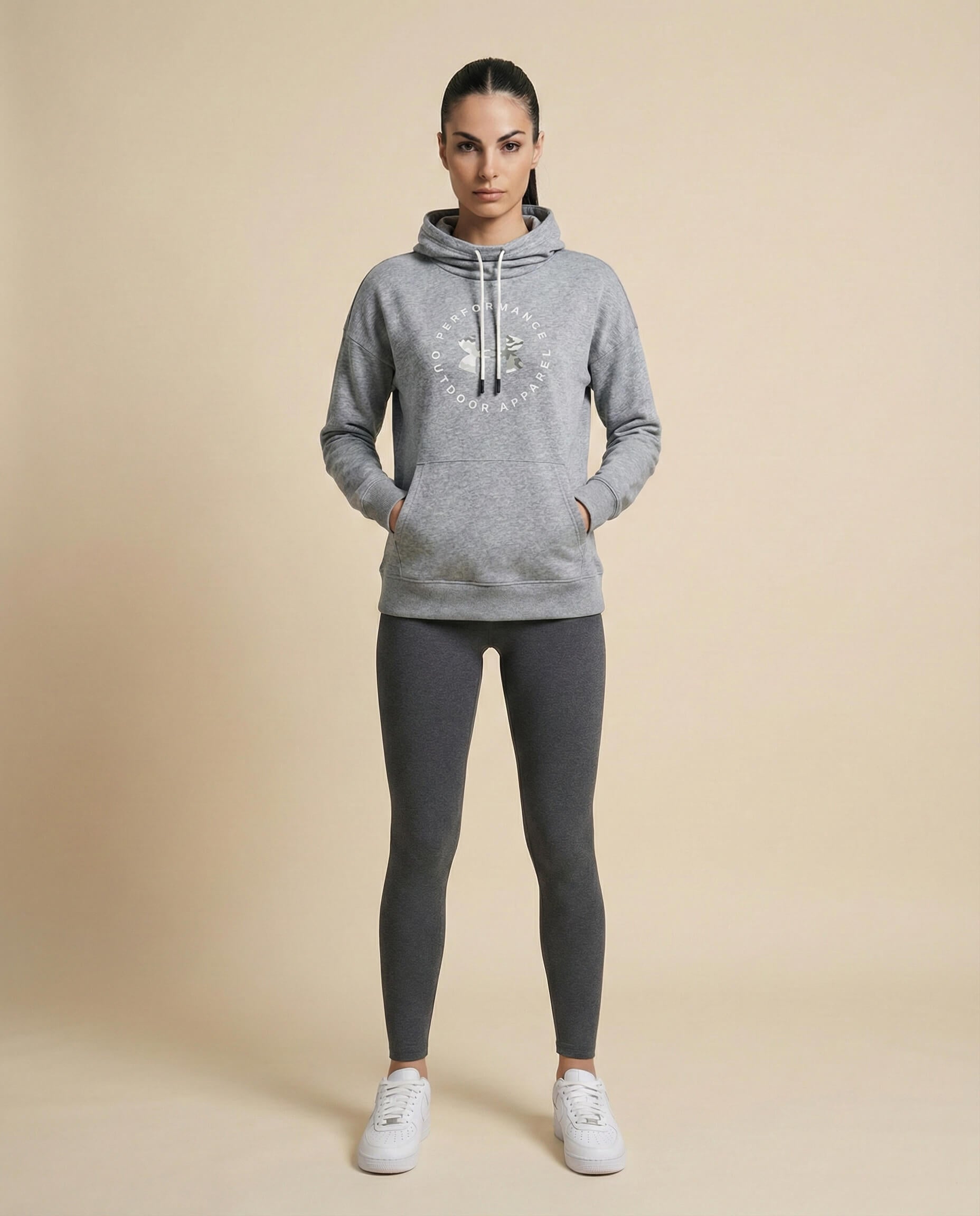 Woman wearing grey hoodie with graphic print, dark leggings, and white sneakers, studio backdrop