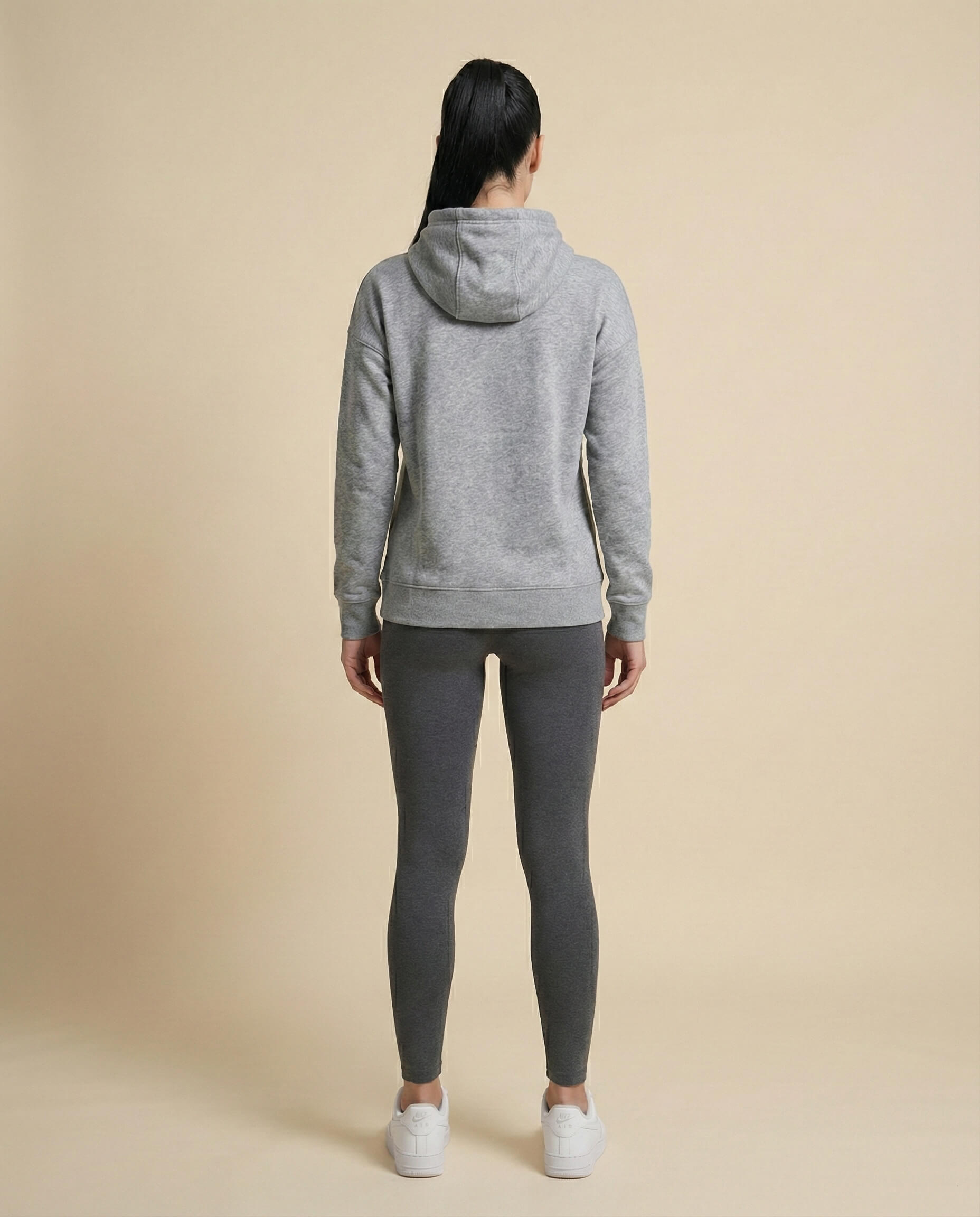 Woman in grey hoodie, dark leggings, and white sneakers standing against beige background