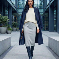Woman in navy coat, cream sweater, grey skirt, and boots walking in modern city plaza
