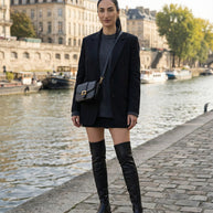 Woman in black blazer, sweater, black mini skirt, and knee-high boots by a riverside