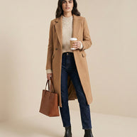 Woman wearing a camel wool coat, cream sweater, jeans, black boots, holding brown tote bag.