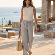Woman in sleeveless ribbed top, light linen pants, sandals and straw bag outdoors by the sea
