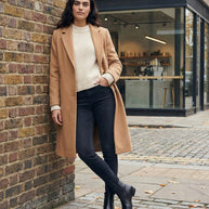 Woman in camel coat, cream sweater, black jeans, and ankle boots standing on city street